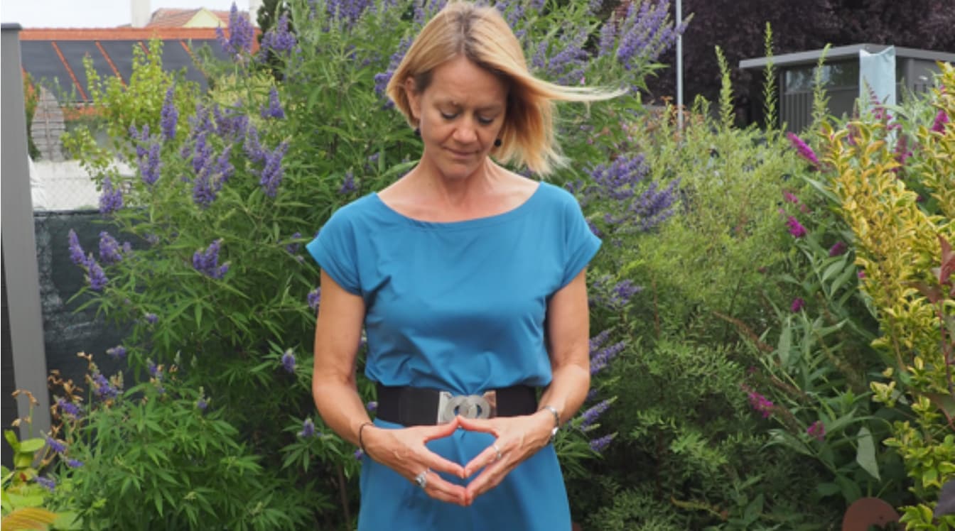 Eine Frau in einem türkis-blau-farbenen Kleid steht in einem blühenden Garten und hält ihre Hände vor dem Körper, wobei sich ihre Fingerspitzen berühren.