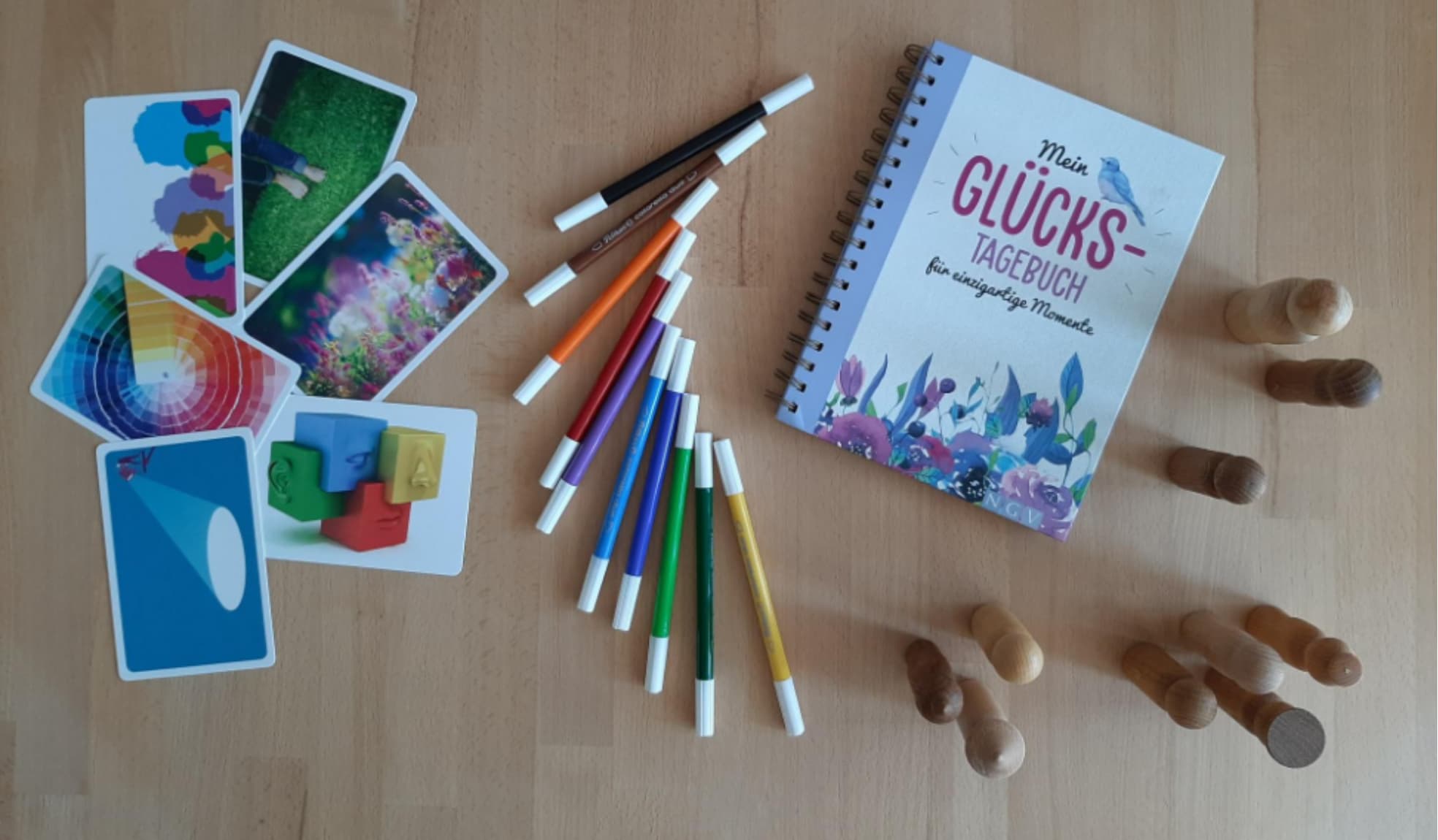 Auf dem Bild sind von links nach rechts durcheinanderliegende bunte Karten, bogenförmig angeordnete bunte Filzstifte, ein liegendes Glückstagebuch sowie Holzfiguren in verschiedenen Positionen zu sehen – als Symbole für kreative Methoden in der psychosozialen Begleitung.