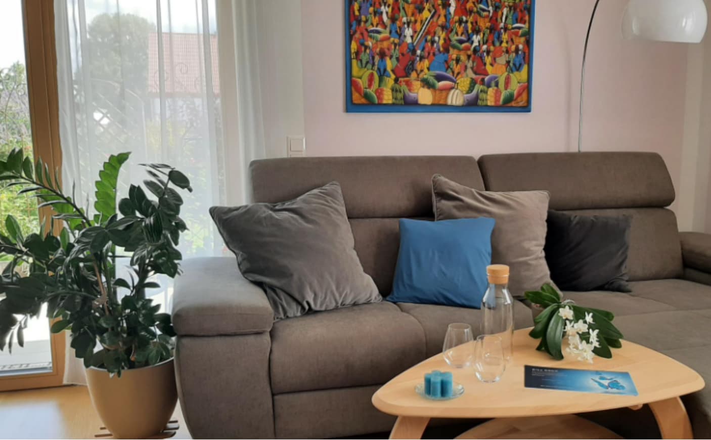 Gemütlicher Beratungsraum mit grauem Sofa, mehreren Kissen und einem hellen Couchtisch mit Wasserflasche, Gläsern, Blumen und einer kleinen Kerze. Links steht eine große Zimmerpflanze, an der Wand hängt ein farbenfrohes Gemälde.