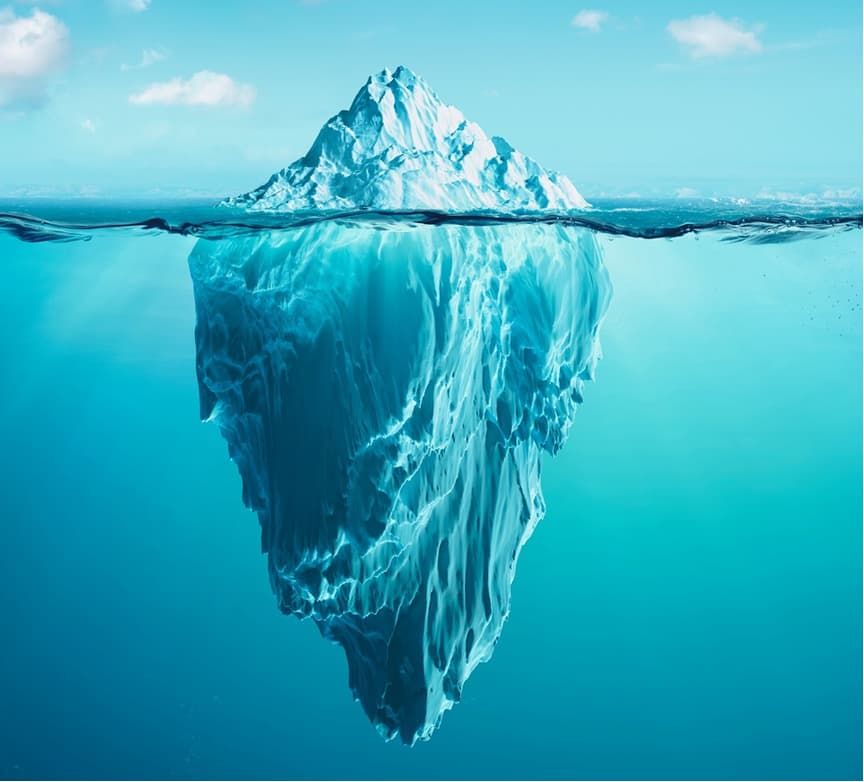 Ein großer Eisberg schwimmt im klaren, blauen Meer; nur die schneebedeckte Spitze ragt über die Wasseroberfläche, während der wesentlich größere, zerklüftete Teil unter Wasser sichtbar ist. Im Hintergrund sind blauer Himmel und einige Wolken zu sehen.