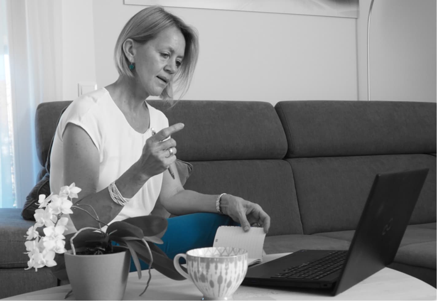 Frau mit Notizblock und Stift auf einer Couch, vertieft in ein Online-Gespräch; auf dem Tisch stehen ein Laptop, eine blühende Orchidee und eine Kaffeetasse.