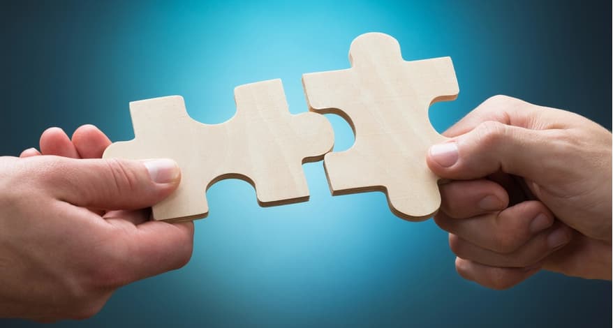 Zwei Hände halten jeweils ein hölzernes Puzzleteil und führen sie zusammen. Die ineinandergreifenden Teile symbolisieren Zusammenarbeit, Verbindung oder das gemeinsame Lösen einer Aufgabe.